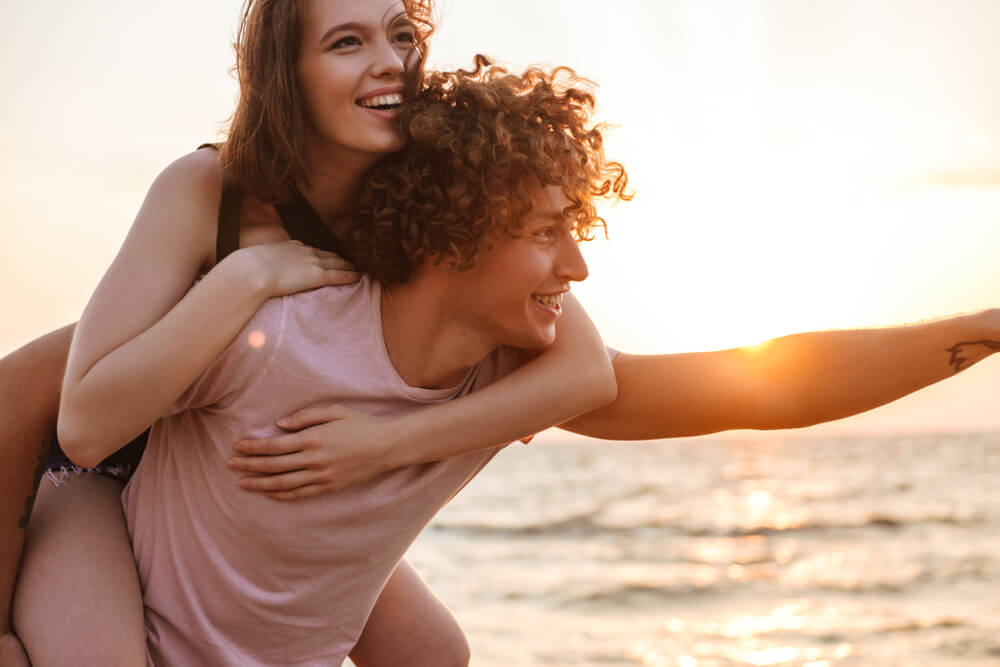 Woman getting a piggy back ride from curly haired on the beach.